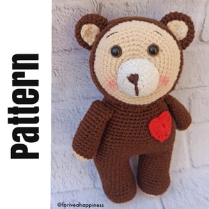 Puede incluir: Un oso de peluche de crochet marrón que lleva un gorro de crochet beige con un corazón blanco en el pecho. El oso tiene ojos marrones y una nariz blanca.