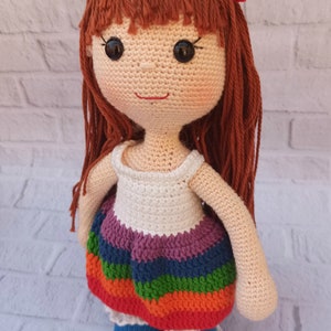 Pu&ograve; includere: Una bambola all'uncinetto con i capelli castani, un vestito bianco con una gonna a righe arcobaleno e scarpe blu. La bambola indossa una fascia per capelli con un fiore.