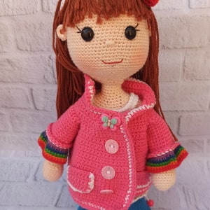 Pu&ograve; includere: Una bambola all'uncinetto con lunghi capelli castani, che indossa una giacca rosa con bordino bianco, jeans blu e una fascia arcobaleno con un fiocco.