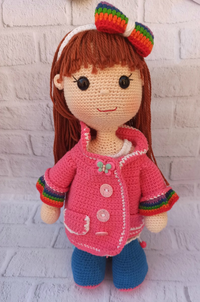 Pu&ograve; includere: Una bambola all'uncinetto con lunghi capelli castani, che indossa una giacca rosa con bordino bianco, jeans blu e una fascia arcobaleno con un fiocco.