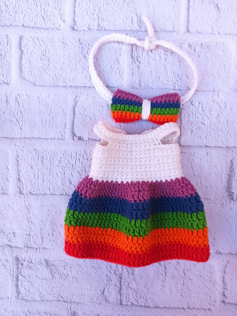 Pu&ograve; includere: Un vestito bianco all'uncinetto con strisce arcobaleno e una fascia per capelli abbinata con un fiocco arcobaleno.