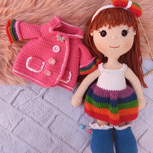 Pu&ograve; includere: Una bambola all'uncinetto con i capelli castani che indossa un vestito a righe arcobaleno e una giacca di uncinetto rosa. La bambola indossa stivali di uncinetto blu.