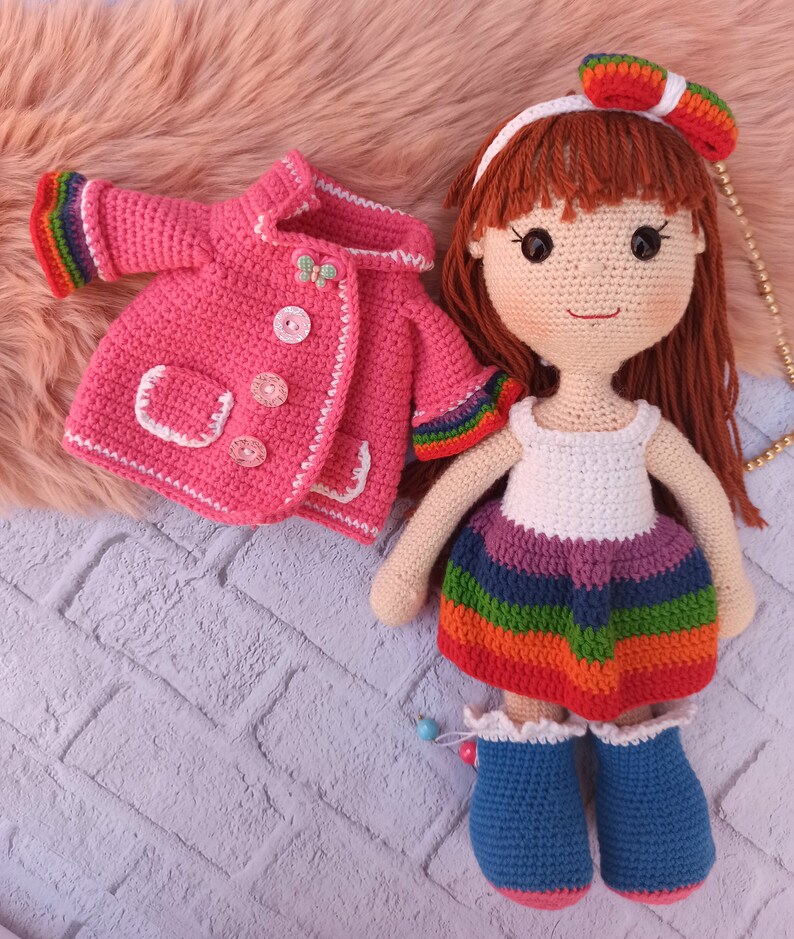 Pu&ograve; includere: Una bambola all'uncinetto con i capelli castani che indossa un vestito a righe arcobaleno e una giacca di uncinetto rosa. La bambola indossa stivali di uncinetto blu.