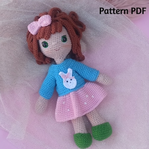Puede incluir: Muñeca de crochet con cabello castaño, un lazo rosa, un suéter azul con una aplicación de conejo blanco y una falda de lunares rosa y blanco. La muñeca lleva zapatos verdes y está acostada sobre una superficie brillante rosa y blanca. El texto "Pattern PDF" es visible en la esquina superior derecha de la imagen.