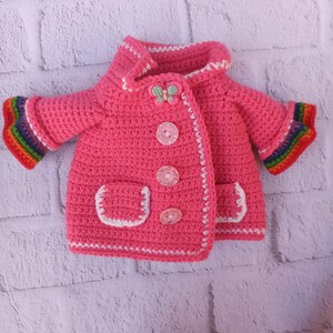 Pu&ograve; includere: Una giacca da beb&eacute; rosa in crochet con bordi bianchi e strisce arcobaleno sulle maniche. La giacca ha una tasca sul davanti ed &egrave; decorata con bottoni e una farfalla.