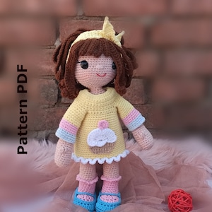 Puede incluir: Muñeca de crochet con cabello castaño, vistiendo un vestido amarillo con un diseño de cupcake, mangas a rayas rosa y azul, calcetines rosas y zapatos azules. La muñeca está de pie sobre una superficie peluda rosa y marrón.