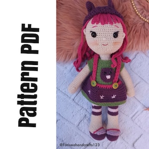 Puede incluir: Patrón de muñeca de crochet en PDF. La muñeca lleva un vestido verde y morado con una diadema de rayas rosa y blancas. La muñeca tiene el pelo de lana rosa y los ojos marrones.