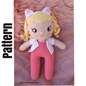 Könnte beinhalten: Gehäkelte Puppe mit blonden Haaren, die einen rosafarbenen Overall und eine weiße Strickjacke trägt. Die Puppe hat einen süßen Ausdruck mit großen Augen und einem Lächeln. Die Puppe trägt Katzenohren.