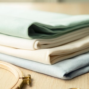 Op de afbeelding: Vier stukken witte Aida-stof gevouwen op een houten oppervlak. De stof is gevouwen in een stapel met het bovenste stuk lichtgroen. Een houten borduurring en een gouden borduurschaar liggen ook op het oppervlak.