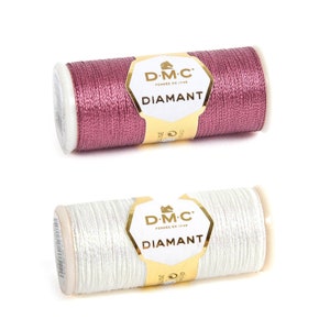 Könnte beinhalten: Zwei Spulen DMC Diamant Metallic-Stickgarn. Eine Spule ist rosa und die andere ist weiß.
