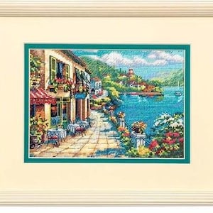 Könnte beinhalten: Ein Kreuzstichmuster, das ein farbenfrohes Küstendorf mit einem Restaurant, einem Café und Blumen zeigt. Die Szene zeigt ein blaues Meer, ein weißes Gebäude mit einer roten Markise und eine gepflasterte Straße.