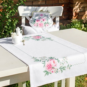 May include: White table runner with a delicate floral embroidery design featuring pink roses and green leaves. The runner has a scalloped edge and is displayed on a white table with a white chair and a small white pitcher.
