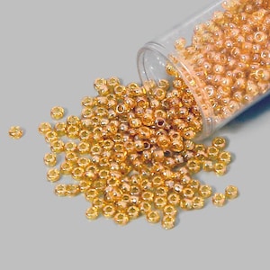 Könnte beinhalten: Ein klares Glasgefäß gefüllt mit kleinen, runden, goldbraunen Perlen. Die Perlen fallen aus dem Gefäß auf eine graue Oberfläche.