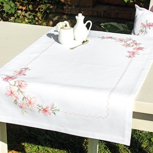 May include: White table runner with a pink floral embroidery design. The runner is on a white table with a white pitcher and a white cup on the table.