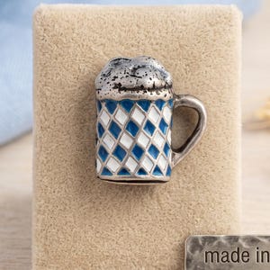 Puede incluir: Un broche plateado con forma de jarra de cerveza con un patrón de rombos azules y blancos y una espuma texturizada. Una pequeña etiqueta metálica rectangular junto al broche muestra el texto: made in Austria, real silver plated.