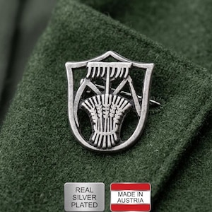 Könnte beinhalten: Versilberte Brosche mit Schilddesign, auf grünem Stoff befestigt. Die Brosche hat ein kompliziertes Muster im Schild. Das Bild enthält auch zwei kleine rechteckige Etiketten, eines mit der Aufschrift "REAL SILVER PLATED" und das andere mit der österreichischen Flagge und "MADE IN AUSTRIA."