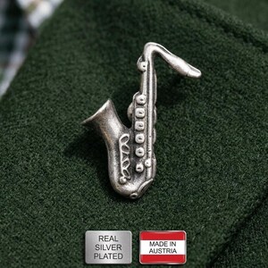 Könnte beinhalten: Ein silberbeschlagener Saxophon-Anstecker, präsentiert auf einem dunkelgrünen Stoff. Der Anstecker ist detailliert mit Klappen und anderen Merkmalen eines Saxophons versehen. Das Bild enthält auch zwei kleine rechteckige Etiketten, eines mit der Aufschrift "REAL SILVER PLATED" und das andere "MADE IN AUSTRIA".