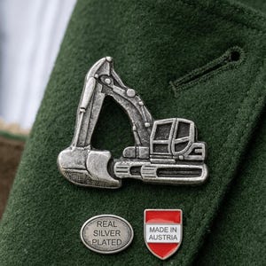 Könnte beinhalten: Ein silberfarbener Bagger-Anstecker und zwei kleinere Anstecknadeln sind an einer dunkelgrünen Jacke befestigt. Eine ovale Nadel trägt die Aufschrift "REAL SILVER PLATED". Die andere ist ein rot-weißes Schild mit der Aufschrift "MADE IN AUSTRIA".