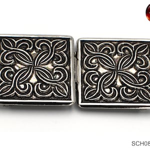May include: A pair of silver-toned metal belt buckles with an intricate black floral design. The buckles are square shaped and have a raised design.