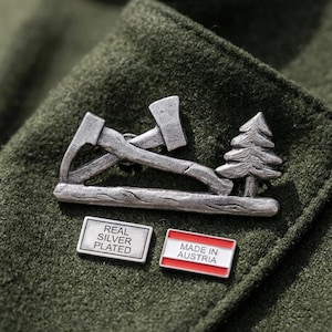 Puede incluir: Un broche plateado con dos hachas cruzadas sobre un tronco junto a un pino. Debajo del broche hay dos pequeños pines rectangulares; uno dice REAL SILVER PLATED y el otro MADE IN AUSTRIA con franjas rojas y blancas.