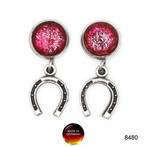 Può includere: Un paio di orecchini argentati. Ogni orecchino presenta una parte superiore rotonda, rossa e rosa piena di glitter e un ciondolo a forma di ferro di cavallo. Chiusura a perno. L'immagine include anche un logo "Made in Germany" e il numero "8480".