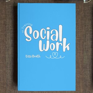 Puede incluir: Un libro azul claro con el texto "In Training Social Work" en fuente cursiva blanca. El nombre del autor, "Lisa Smith", está impreso debajo del título. Un pequeño corazón con un diseño de espiral está debajo del nombre del autor.