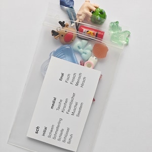 May include: A transparent plastic bag containing various miniature figurines and charms. The bag includes a small reindeer, a green frog, a tiny person, and other assorted shapes. A white card with German words is visible.