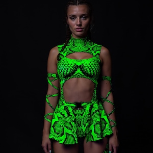 Peut inclure: Une tenue néon verte à motif peau de serpent. La tenue comprend un haut court à col montant avec une découpe en trou de serrure, un short assorti et des manchettes. L'ensemble est posé sur un fond noir, soulignant la couleur et le design vibrants.