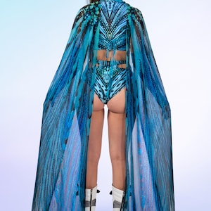 May include: A two-piece swimsuit featuring a blue and black butterfly wing pattern, paired with a matching sheer floor-length cape that drapes from the shoulders. The set is worn with white platform boots featuring black vertical zipper details.
