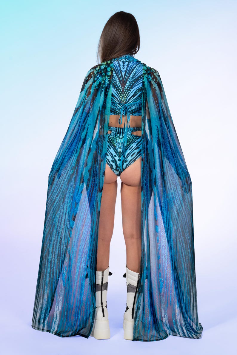 May include: A two-piece swimsuit featuring a blue and black butterfly wing pattern, paired with a matching sheer floor-length cape that drapes from the shoulders. The set is worn with white platform boots featuring black vertical zipper details.