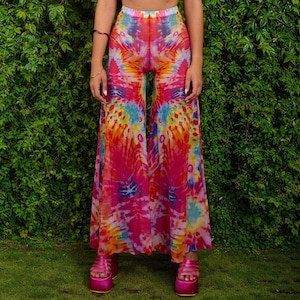 May include: Wide-leg pants with a vibrant tie-dye pattern in shades of pink, orange, yellow, and blue. The pants are made of a sheer fabric and are paired with platform sandals. The background features lush green foliage.