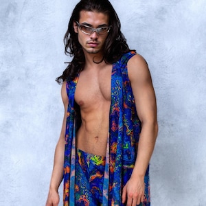 Op de afbeelding: Een mouwloos, openvallend kledingstuk en bijpassende shorts in een levendig blauw, oranje en groen abstract patroon. De outfit wordt gedragen door een persoon met lang donker haar en een zilverkleurige bril.