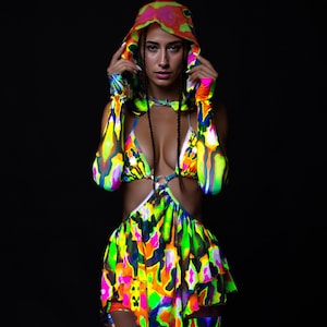 May include: A brightly coloured outfit with a hood, bikini top, and skirt. The outfit has an abstract pattern in neon shades of green, yellow, pink, and blue. The model is wearing the outfit against a black background.