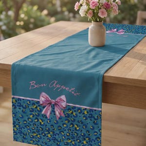 Può includere: Un runner da tavola color verde acqua con un motivo leopardato blu e giallo alle estremità, accentuato da un fiocco rosa e dalla scritta Bon Appetit in corsivo rosa. Un piccolo vaso beige con garofani rosa e bianchi è posto sul runner.