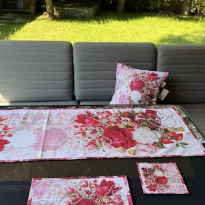 Puede incluir: Un camino de mesa rectangular, un mantel individual cuadrado, un posavasos cuadrado pequeño y una funda de cojín cuadrada, todos con fondo blanco y un estampado floral vibrante de rosas rojas, tulipanes, flores blancas, granadas y collares de perlas.