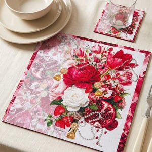 Puede incluir: Un mantel individual cuadrado y un posavasos a juego con un estampado floral vibrante de tulipanes rojos, rosas blancas, granadas, fresas y collares de perlas. El diseño incluye el texto LaMua en la esquina inferior izquierda del mantel.