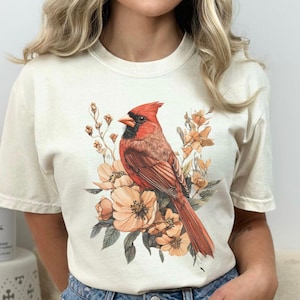 May include: A white t-shirt with a red cardinal and floral design. The shirt has a crew neck and short sleeves. The text "IVORY" is visible in the lower left corner of the image.
