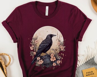 May include: A maroon t-shirt with a graphic of a black crow perched on a rock with pink flowers and a full moon in the background.
