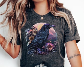May include: A dark gray t-shirt with a graphic print of a black raven perched on a branch with purple and pink flowers. The raven is surrounded by a sunset-colored circle with a mountain range in the background.