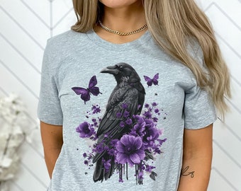 May include: A gray t-shirt with a black raven and purple flowers printed on the front. The raven is perched on a branch of flowers and has its wings spread out. There are two butterflies on either side of the raven.