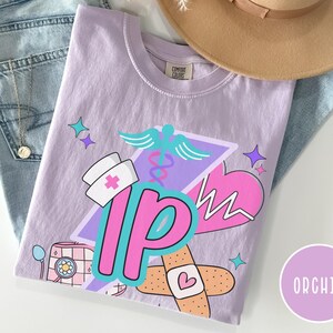 May include: Orchid-colored t-shirt with a graphic design featuring a nurse's hat, a caduceus, a heart, and the letters "IP". The design includes a bandage with a heart and a retro-style medical device. The shirt is folded on top of blue jeans.