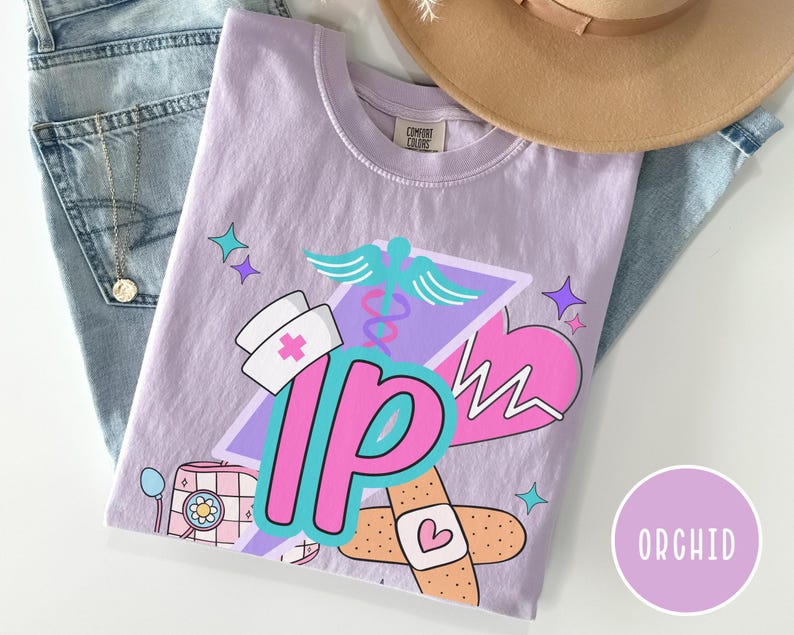 May include: Orchid-colored t-shirt with a graphic design featuring a nurse's hat, a caduceus, a heart, and the letters "IP". The design includes a bandage with a heart and a retro-style medical device. The shirt is folded on top of blue jeans.