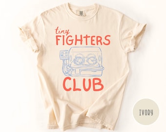 以下が含まれることがあります： 「tiny FIGHTERS CLUB」の赤い文字と、インキュベーターの青いイラストが描かれた、アイボリー色のTシャツ。クルーネックで袖はロールアップされています。Tシャツの色はアイボリーです。