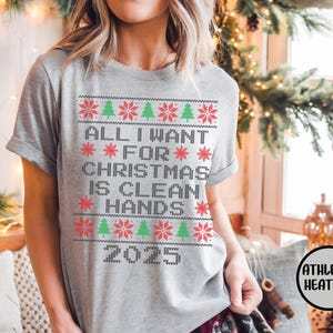 May include: Athletic heather gray t-shirt with a festive Christmas design. The shirt features the text "ALL I WANT FOR CHRISTMAS IS CLEAN HANDS 2025" in a pixelated style with red snowflakes and green Christmas trees. The shirt is short-sleeved.