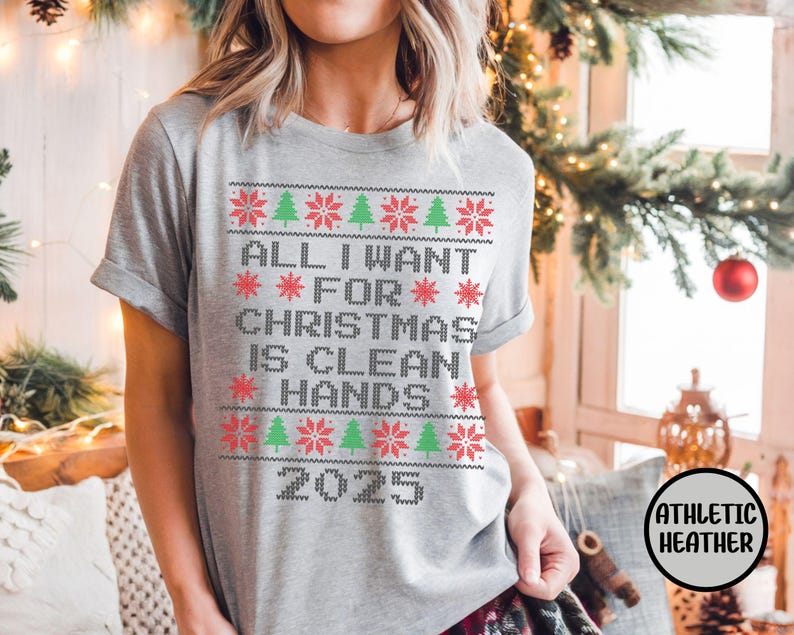 May include: Athletic heather gray t-shirt with a festive Christmas design. The shirt features the text "ALL I WANT FOR CHRISTMAS IS CLEAN HANDS 2025" in a pixelated style with red snowflakes and green Christmas trees. The shirt is short-sleeved.