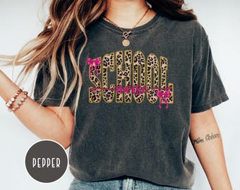 Pode incluir: Uma t-shirt de manga curta cinzento-carvão com o texto SCHOOL NURSE em padrão leopardo e contornos dourados. A palavra SCHOOL está em letras grandes e a palavra nurse em letra cursiva rosa com dois laços cor-de-rosa.
