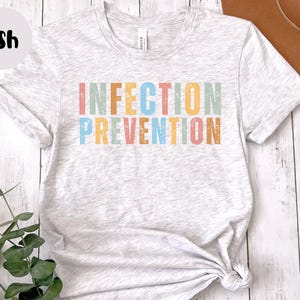 May include: Light gray t-shirt with the words "INFECTION PREVENTION" in colorful, block letters. The shirt is knotted at the bottom and has rolled-up sleeves. The word "Ash" is in a black circle in the upper left corner.