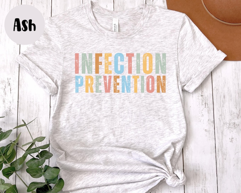 May include: Light gray t-shirt with the words "INFECTION PREVENTION" in colorful, block letters. The shirt is knotted at the bottom and has rolled-up sleeves. The word "Ash" is in a black circle in the upper left corner.