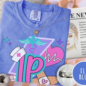 May include: A blue t-shirt with a graphic design featuring a nurse's hat, a medical symbol, and the letters "IP." The design includes a pink heart and band-aids. A silver necklace with a pendant is draped across the shirt.