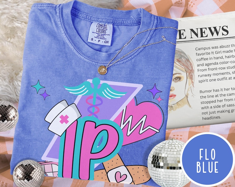 May include: A blue t-shirt with a graphic design featuring a nurse's hat, a medical symbol, and the letters "IP." The design includes a pink heart and band-aids. A silver necklace with a pendant is draped across the shirt.